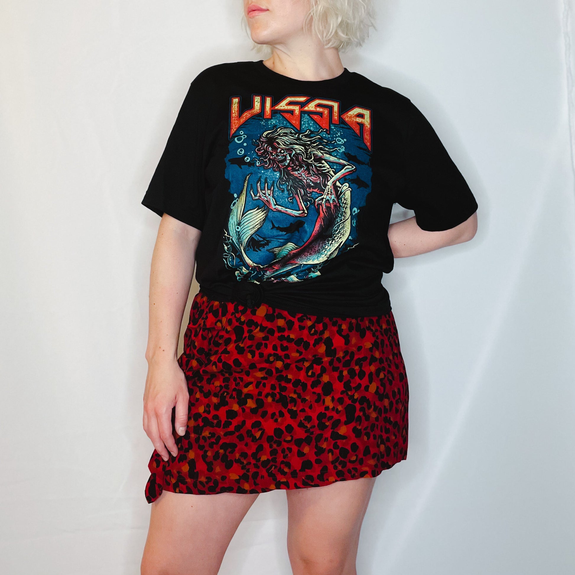 Blonde woman is wearing a black metal band style t-shirt and red leopard print skirt; the t-shirt shows an illustration of a skeleton siren under the sea and "VISSIA" across the top