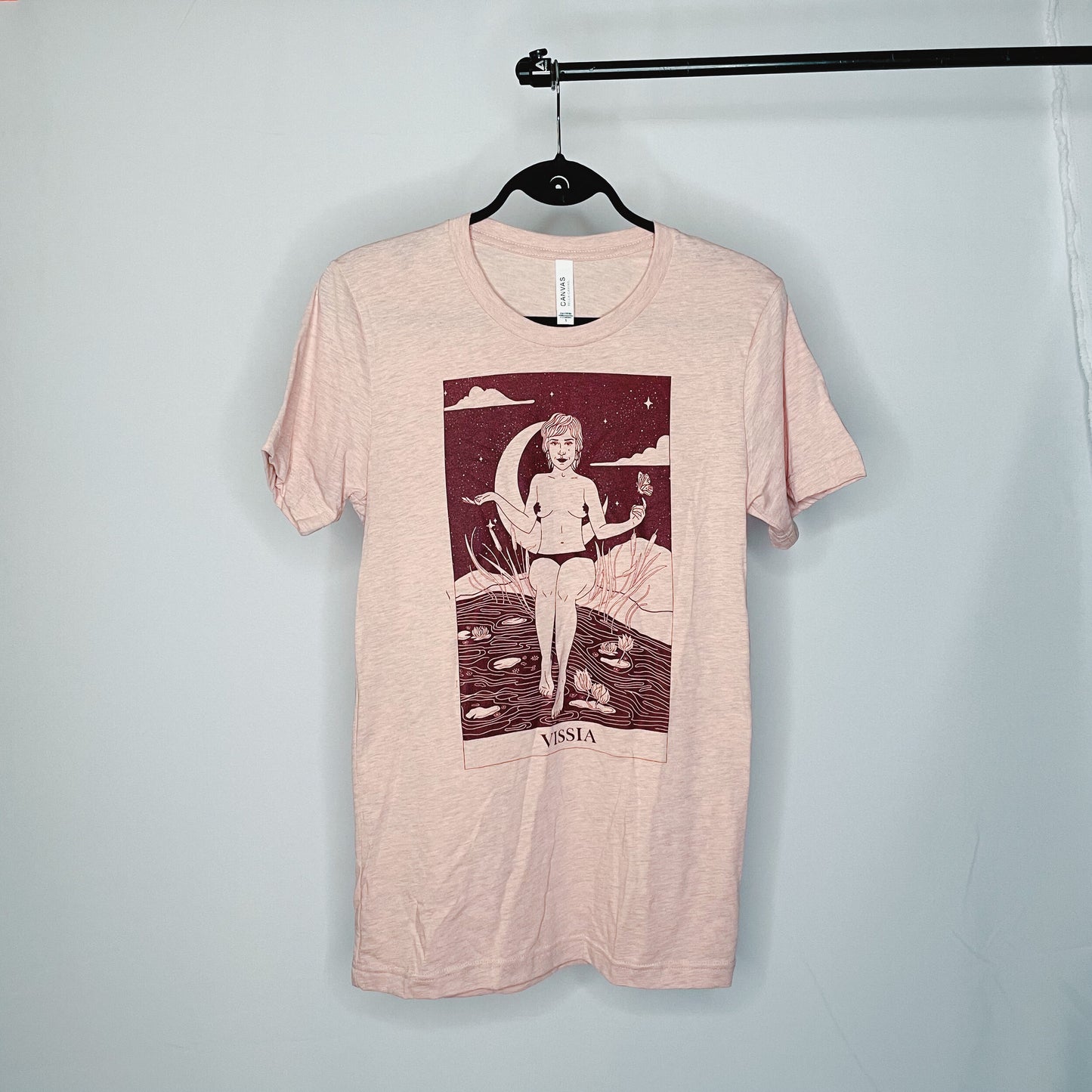 heather peach t-shirt with rusty red illustration; the illustration is in the style of a tarot card and shows an unclothed woman sitting on the banks of a river; a butterfly hovers above her left hand and her right hand is extended; a crescent moon hangs against a dark starry sky