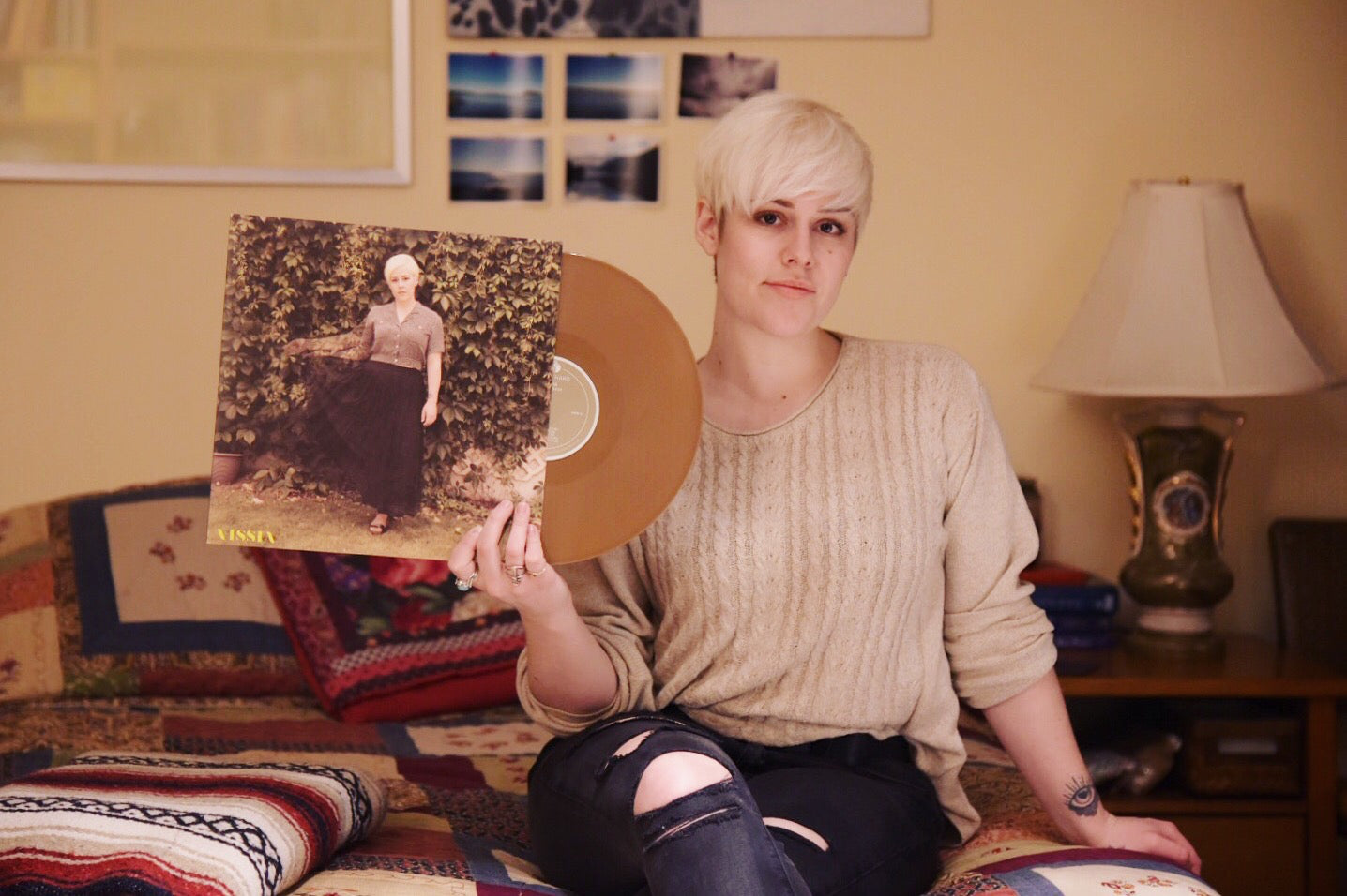 Woman with short blonde hair is wearing an oatmeal coloured sweater and distressed black jeans; she is sitting on a patchwork quilted bed and holding a vinyl record in her right hand; the cover shows a short haired blonde woman in long black skirt and grey button-up shirt posing against a wall of green vines